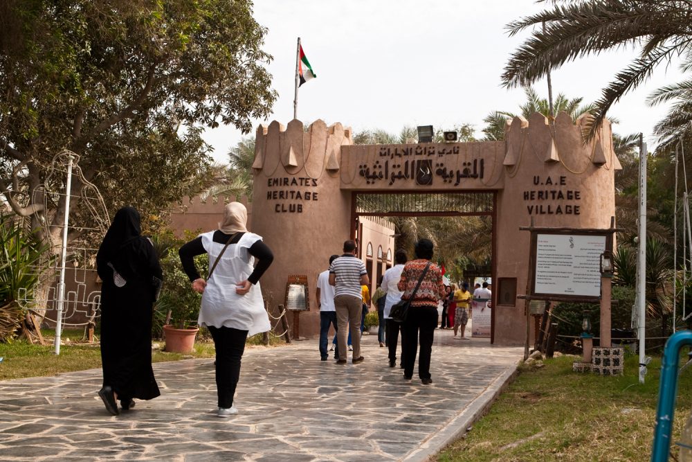 People walking towards the entrance of the UAE Heritage Village with a flag on top.